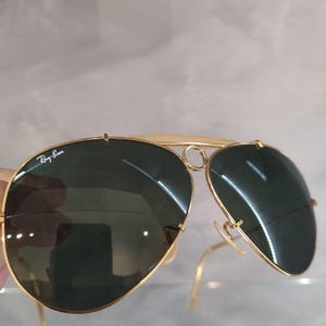 Puede incluir: Gafas de sol aviador Ray-Ban doradas con lentes verdes. Las gafas tienen un puente doble y un marco de metal.