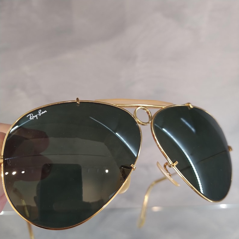 Puede incluir: Gafas de sol aviador Ray-Ban doradas con lentes verdes. Las gafas tienen un puente doble y un marco de metal.