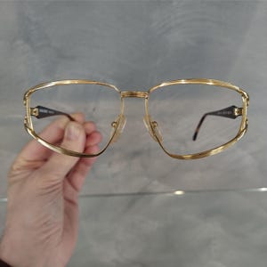 Pu&ograve; includere: Occhiali da vista in metallo dorato con montatura quadrata e lenti trasparenti. Gli occhiali hanno una montatura a doppio strato.
