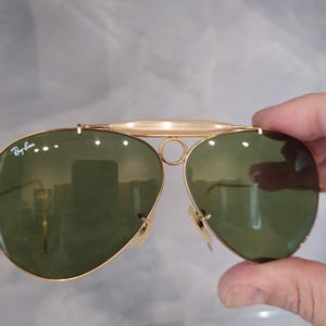 May include: Gold-tone aviator sunglasses with green lenses and a Ray-Ban logo on the left lens.