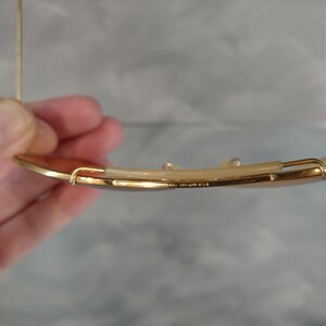 May include: Gold-toned sunglasses with a brown lens. The frame has a thin, delicate design. The text "MADE IN U.S.A." is visible on the frame. The sunglasses are held by a person.