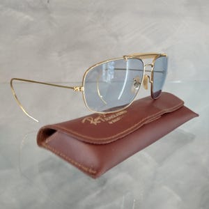 Puede incluir: Gafas de sol aviador doradas con lentes azules y una funda de cuero marr&oacute;n. La funda tiene el texto "Ray-Ban Sunglasses by Bausch & Lomb" impreso en ella.