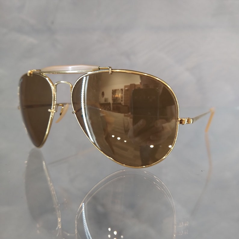May include: Gold aviator sunglasses with brown mirrored lenses. The sunglasses have a thin metal frame and a bridge that connects the two lenses.
