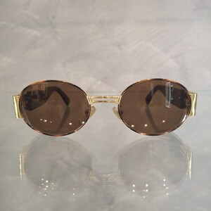 K&ouml;nnte beinhalten: Sonnenbrille mit goldfarbenem Rahmen und braunen Gl&auml;sern. Die runden Gl&auml;ser sind in einem goldenen Rahmen mit Steg und B&uuml;geln eingefasst. Die Sonnenbrille hat einen klassischen, Retro-Stil.