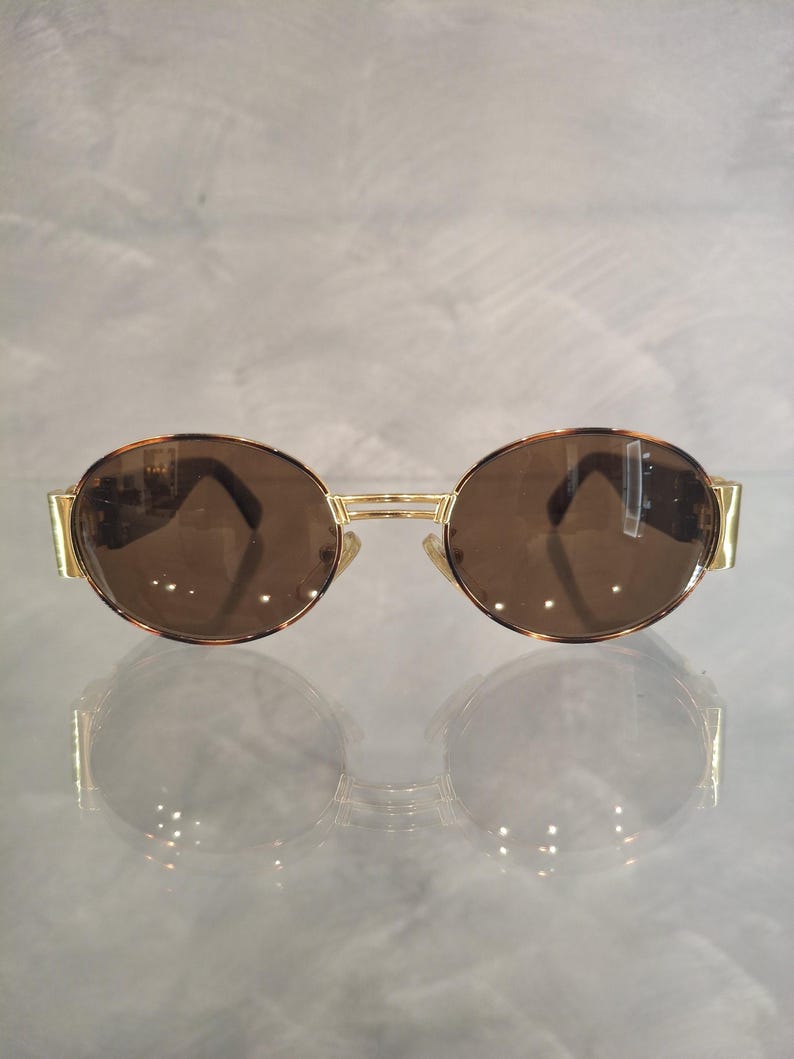 K&ouml;nnte beinhalten: Sonnenbrille mit goldfarbenem Rahmen und braunen Gl&auml;sern. Die runden Gl&auml;ser sind in einem goldenen Rahmen mit Steg und B&uuml;geln eingefasst. Die Sonnenbrille hat einen klassischen, Retro-Stil.