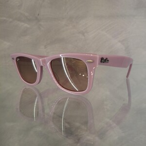 Puede incluir: Gafas de sol rosas con lentes marrones. Las gafas tienen un dise&ntilde;o de montura rectangular cl&aacute;sico. El logotipo de Ray-Ban es visible en la patilla. Las gafas se reflejan en una superficie brillante.