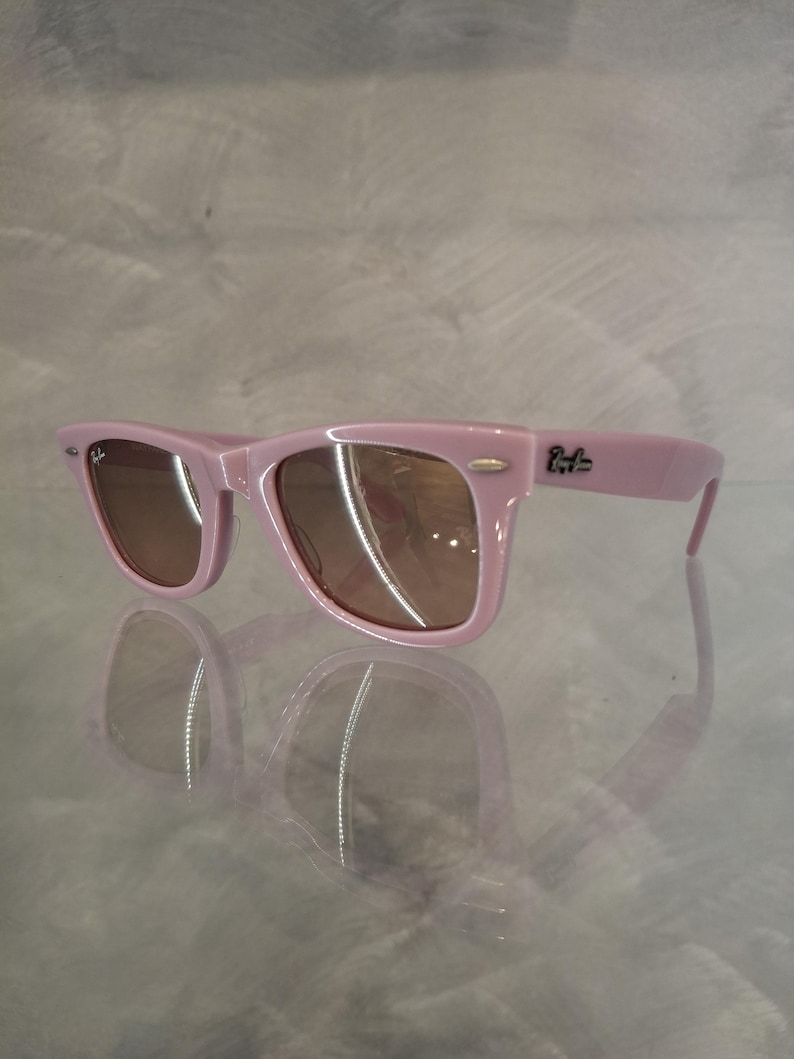 Puede incluir: Gafas de sol rosas con lentes marrones. Las gafas tienen un dise&ntilde;o de montura rectangular cl&aacute;sico. El logotipo de Ray-Ban es visible en la patilla. Las gafas se reflejan en una superficie brillante.