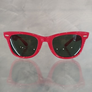 Può includere: Occhiali da sole in plastica rossa in stile wayfarer classico. Gli occhiali hanno lenti scure e un accento dorato sulla montatura.