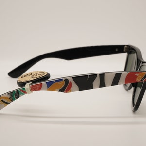 K&ouml;nnte beinhalten: Schwarze Sonnenbrille mit einem wei&szlig;en und schwarzen Zebra-Muster an den B&uuml;geln. Die B&uuml;gel haben auch farbenfrohe abstrakte Designs. Die Sonnenbrille hat gr&uuml;ne Gl&auml;ser.