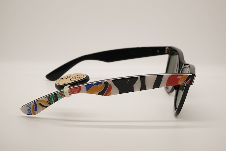 K&ouml;nnte beinhalten: Schwarze Sonnenbrille mit einem wei&szlig;en und schwarzen Zebra-Muster an den B&uuml;geln. Die B&uuml;gel haben auch farbenfrohe abstrakte Designs. Die Sonnenbrille hat gr&uuml;ne Gl&auml;ser.