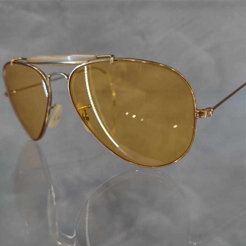 May include: Gold-framed aviator sunglasses with amber-tinted lenses. The sunglasses feature a double bridge and a white brow bar. The lenses are a warm, light brown color, and the frames are a shiny gold.