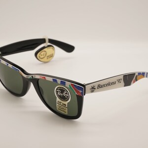 K&ouml;nnte beinhalten: Schwarze Ray-Ban Sonnenbrille mit gr&uuml;nen Gl&auml;sern und einem wei&szlig;en Rahmen mit einem bunten Design und dem Text "Barcelona '92".