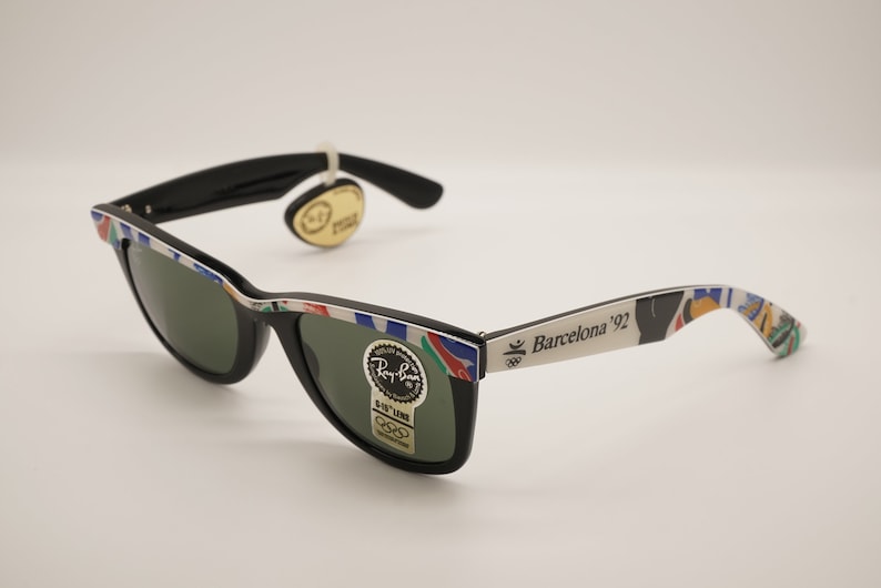 K&ouml;nnte beinhalten: Schwarze Ray-Ban Sonnenbrille mit gr&uuml;nen Gl&auml;sern und einem wei&szlig;en Rahmen mit einem bunten Design und dem Text "Barcelona '92".