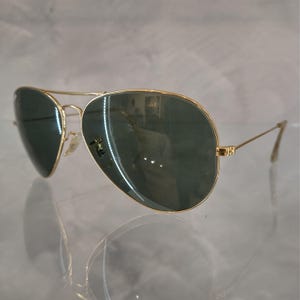 Puede incluir: Gafas de sol aviador doradas con lentes verde oscuro. Las gafas est&aacute;n sobre una superficie blanca.