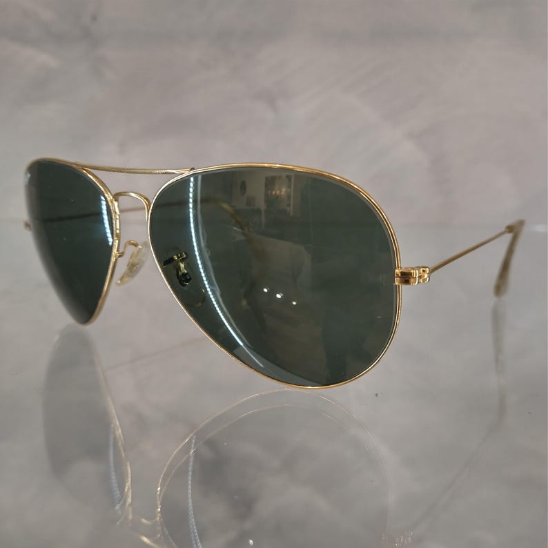 Puede incluir: Gafas de sol aviador doradas con lentes verde oscuro. Las gafas est&aacute;n sobre una superficie blanca.