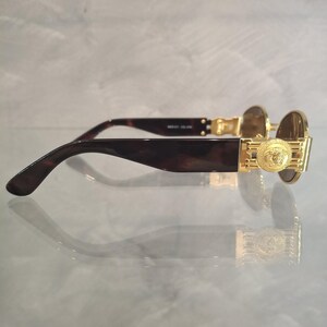 K&ouml;nnte beinhalten: Goldfarbene Sonnenbrille mit einem Medusa-Kopf-Emblem an den B&uuml;geln. Die Fassungen haben ein dunkles Schildpattmuster, und die Gl&auml;ser sind leicht braun get&ouml;nt. Die Sonnenbrille wird auf einer reflektierenden Oberfl&auml;che pr&auml;sentiert.