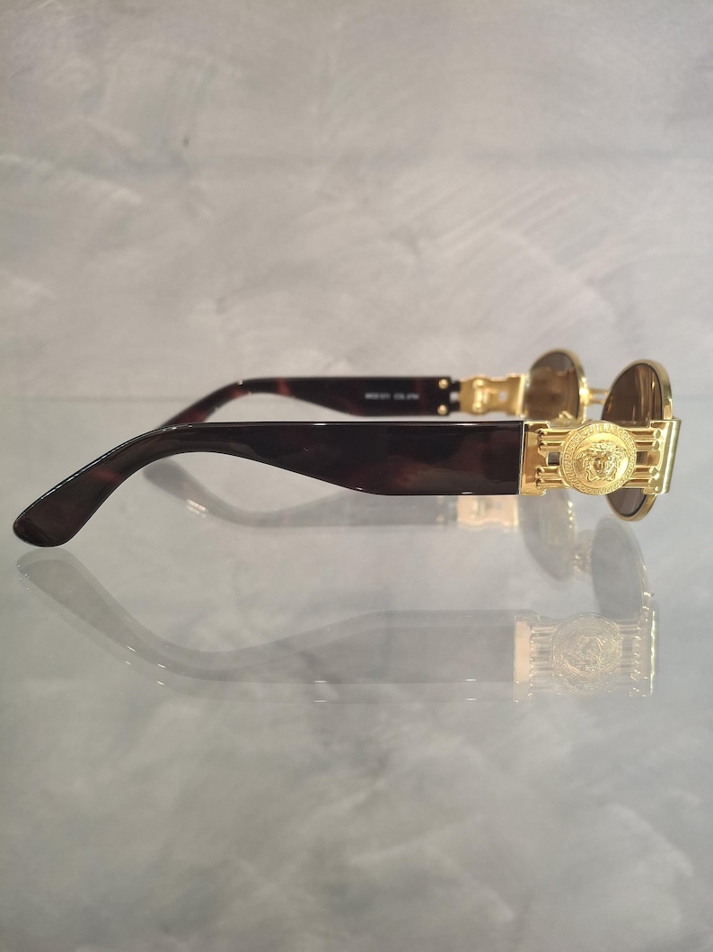 K&ouml;nnte beinhalten: Goldfarbene Sonnenbrille mit einem Medusa-Kopf-Emblem an den B&uuml;geln. Die Fassungen haben ein dunkles Schildpattmuster, und die Gl&auml;ser sind leicht braun get&ouml;nt. Die Sonnenbrille wird auf einer reflektierenden Oberfl&auml;che pr&auml;sentiert.