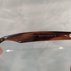 May include: Close-up of the brown tortoiseshell frame of a pair of Wayfarer II sunglasses. The text "WAYFARER II" is engraved on the frame.