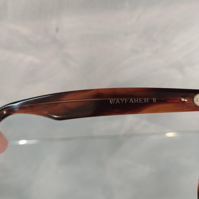 May include: Close-up of the brown tortoiseshell frame of a pair of Wayfarer II sunglasses. The text "WAYFARER II" is engraved on the frame.