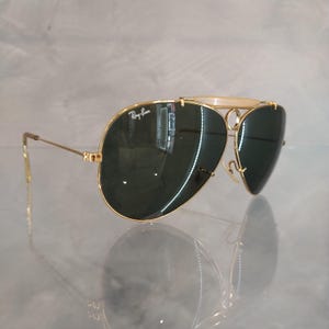 Puede incluir: Gafas de sol aviador doradas con lentes verdes y un logotipo de Ray-Ban en el marco.