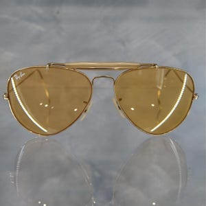 May include: Gold-framed aviator sunglasses with amber-tinted lenses. The sunglasses feature a double bridge and a top bar. The Ray-Ban logo is visible on the lens. The lenses are teardrop-shaped.