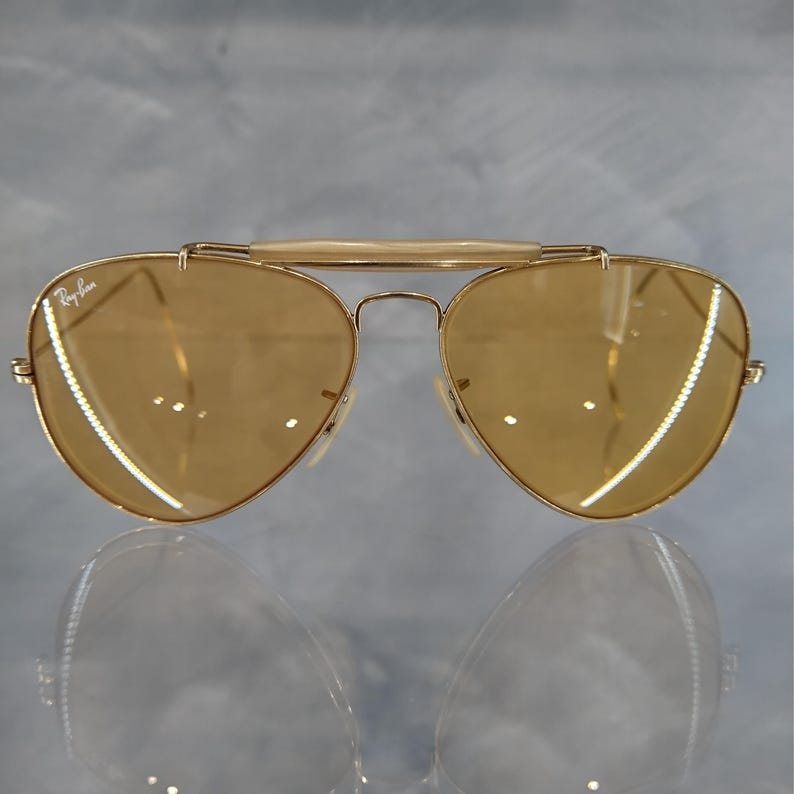 May include: Gold-framed aviator sunglasses with amber-tinted lenses. The sunglasses feature a double bridge and a top bar. The Ray-Ban logo is visible on the lens. The lenses are teardrop-shaped.
