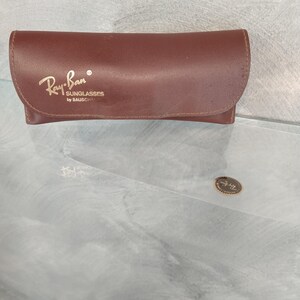 Puede incluir: Una funda de cuero marr&oacute;n para gafas de sol Ray-Ban. La funda tiene el logotipo de Ray-Ban y el texto "Sunglasses by Bausch & Lomb" grabado en la parte delantera.