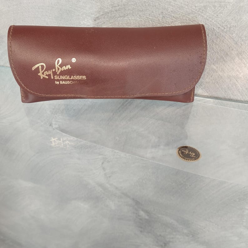 Puede incluir: Una funda de cuero marr&oacute;n para gafas de sol Ray-Ban. La funda tiene el logotipo de Ray-Ban y el texto "Sunglasses by Bausch & Lomb" grabado en la parte delantera.