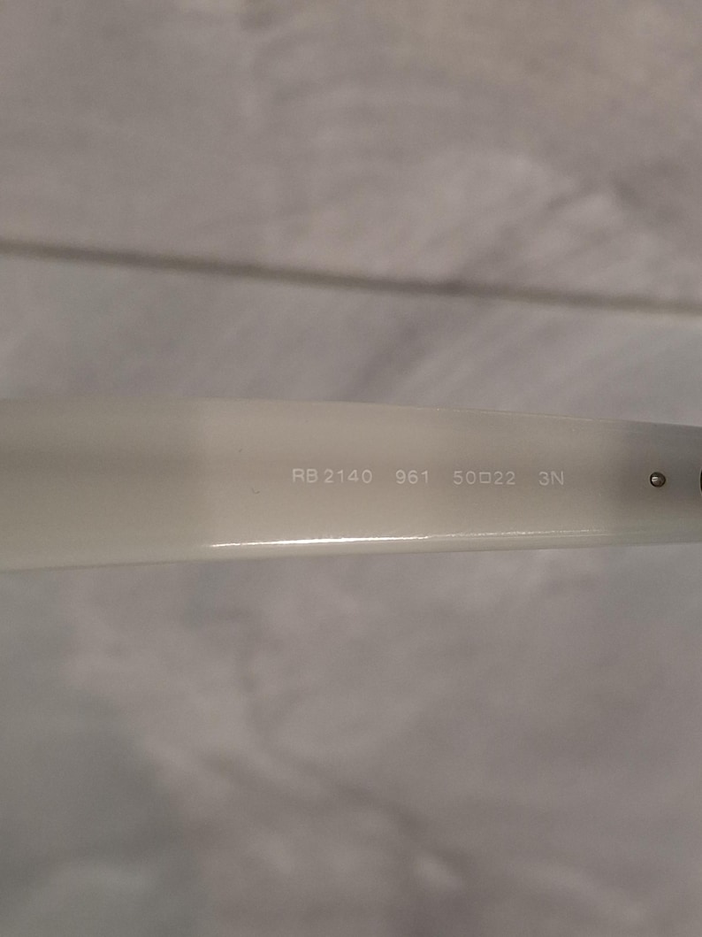 Pu&ograve; includere: Primo piano del braccio di un paio di occhiali da sole bianco sporco. Il testo "RB 2140 961 50▢22 3N" &egrave; stampato sul braccio. Gli occhiali da sole sono di stile classico.