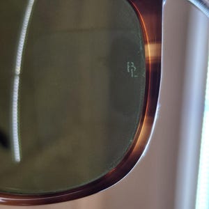 May include: Close-up of sunglasses with a tortoiseshell frame and green lenses. The frame has a glossy finish with a gradient of brown and amber hues. The lenses have a subtle green tint, and the brand logo is visible on the lens.
