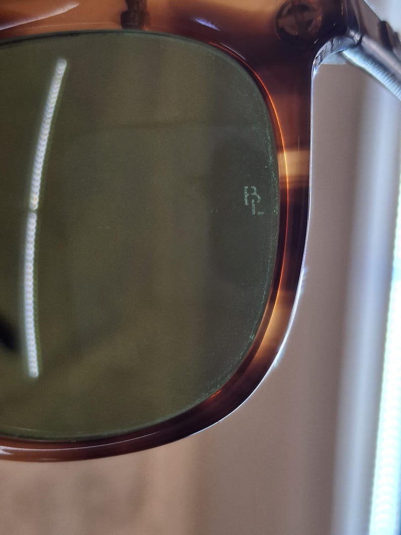 May include: Close-up of sunglasses with a tortoiseshell frame and green lenses. The frame has a glossy finish with a gradient of brown and amber hues. The lenses have a subtle green tint, and the brand logo is visible on the lens.