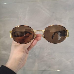 K&ouml;nnte beinhalten: Runde Sonnenbrille mit braunen Gl&auml;sern und einem goldfarbenen Metallrahmen. Die Sonnenbrille hat ein klassisches Design mit schmalem Steg und B&uuml;geln. Die Gl&auml;ser reflektieren die Umgebung.