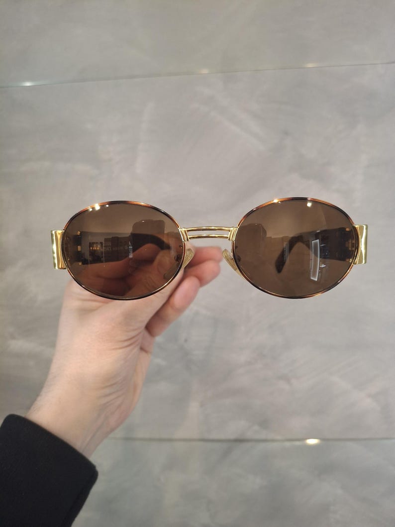 K&ouml;nnte beinhalten: Runde Sonnenbrille mit braunen Gl&auml;sern und einem goldfarbenen Metallrahmen. Die Sonnenbrille hat ein klassisches Design mit schmalem Steg und B&uuml;geln. Die Gl&auml;ser reflektieren die Umgebung.