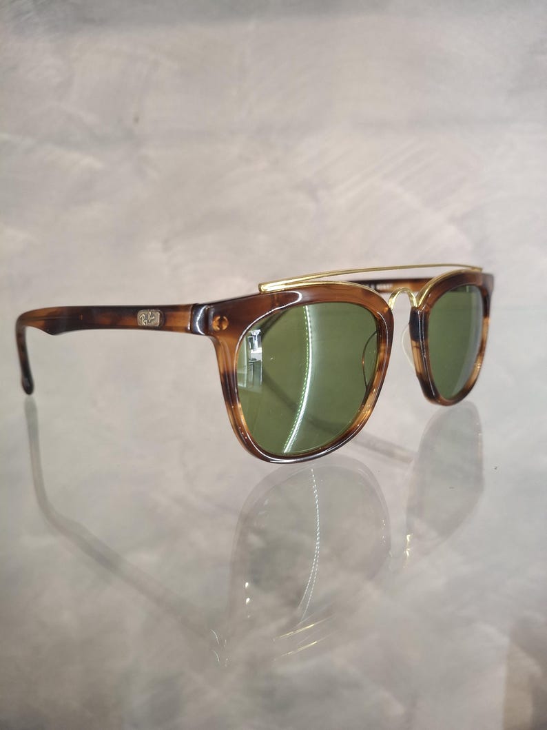 May include: Sunglasses with a brown tortoiseshell frame and green lenses. The sunglasses have a gold-colored metal bridge and arms. The lenses are a dark green color, and the frame has a classic, retro design.