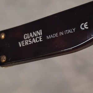 K&ouml;nnte beinhalten: Nahaufnahme eines Teils einer Sonnenbrille. Der Rahmen hat ein dunkles Schildpattmuster. Der Text "GIANNI VERSACE MADE IN ITALY" ist wei&szlig; gedruckt. Das CE-Zeichen ist ebenfalls sichtbar.