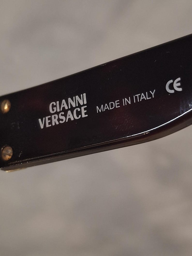 K&ouml;nnte beinhalten: Nahaufnahme eines Teils einer Sonnenbrille. Der Rahmen hat ein dunkles Schildpattmuster. Der Text "GIANNI VERSACE MADE IN ITALY" ist wei&szlig; gedruckt. Das CE-Zeichen ist ebenfalls sichtbar.