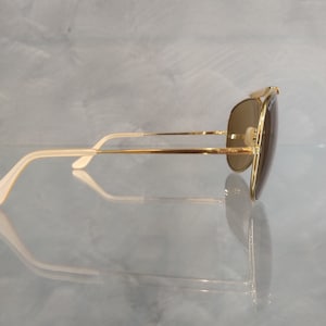 May include: Gold aviator sunglasses with brown lenses and white temple tips. The sunglasses are on a reflective surface.