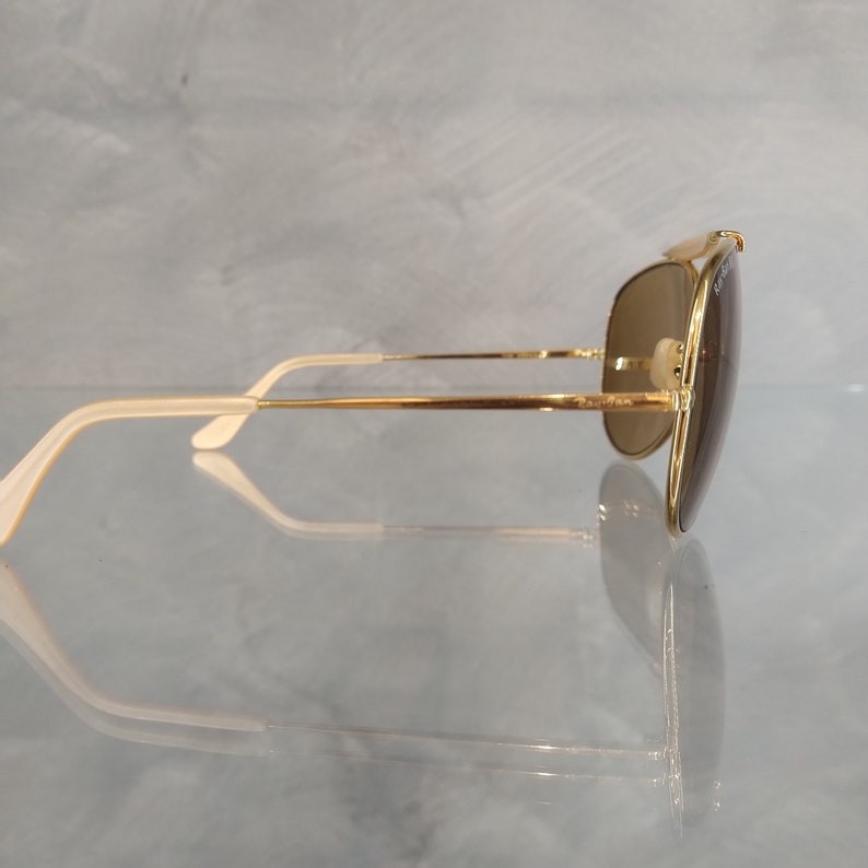 May include: Gold aviator sunglasses with brown lenses and white temple tips. The sunglasses are on a reflective surface.