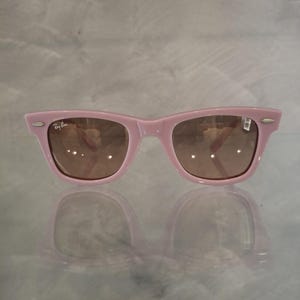 Puede incluir: Gafas de sol rosas estilo Wayfarer con lentes marrones. El logotipo de Ray-Ban es visible en la esquina superior izquierda. Las gafas se reflejan en una superficie brillante.