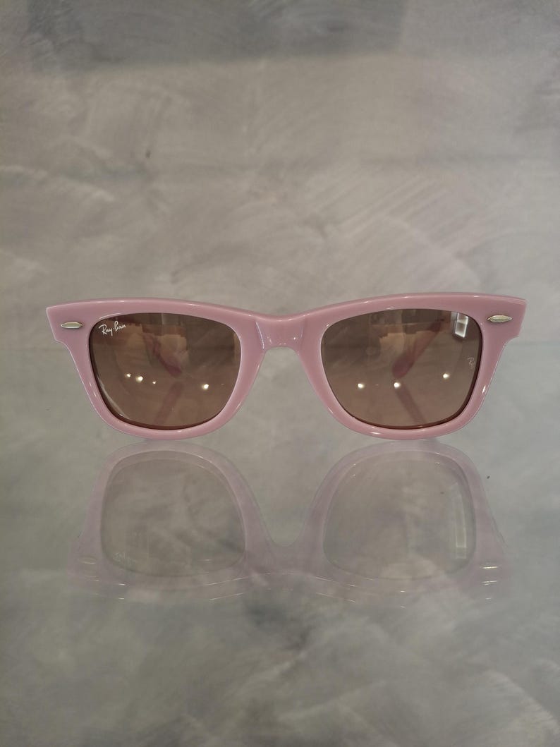 Puede incluir: Gafas de sol rosas estilo Wayfarer con lentes marrones. El logotipo de Ray-Ban es visible en la esquina superior izquierda. Las gafas se reflejan en una superficie brillante.