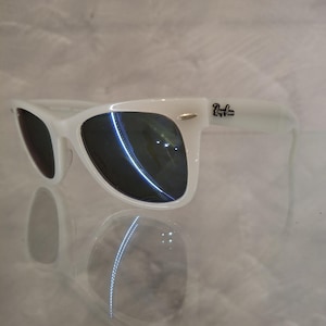 Pu&ograve; includere: Occhiali da sole bianchi con lenti scure. Gli occhiali da sole hanno un classico stile Wayfarer con montatura bianca e il logo Ray-Ban sull'asta. Le lenti riflettono la luce, su sfondo chiaro.