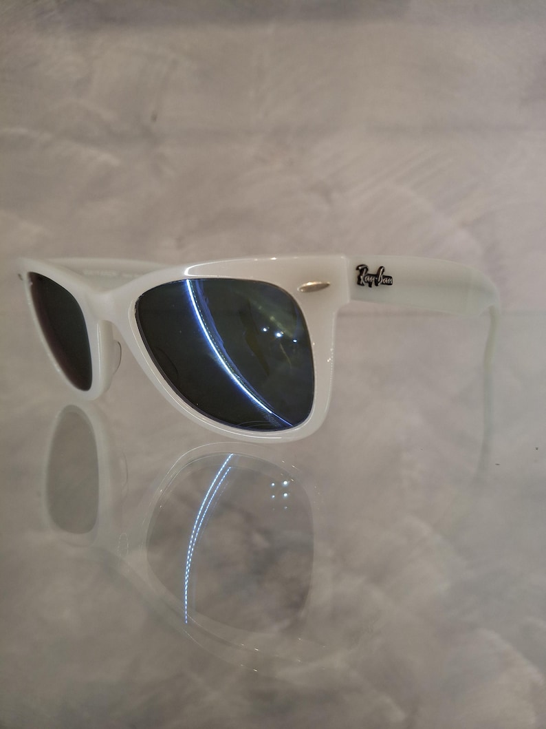 Pu&ograve; includere: Occhiali da sole bianchi con lenti scure. Gli occhiali da sole hanno un classico stile Wayfarer con montatura bianca e il logo Ray-Ban sull'asta. Le lenti riflettono la luce, su sfondo chiaro.