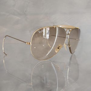 May include: Gold-tone aviator sunglasses with brown gradient lenses and a white bridge. The sunglasses have a Ray-Ban logo on the left lens.