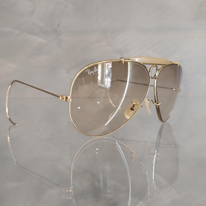 May include: Gold-tone aviator sunglasses with brown gradient lenses and a white bridge. The sunglasses have a Ray-Ban logo on the left lens.