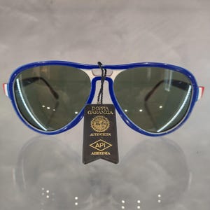 May include: Blue and white aviator sunglasses with green lenses. The sunglasses have a tag attached that says "Doppia Garanzia" and "Ray-Ban" with a logo. The tag also says "Autenticita" and "API Assistenza".