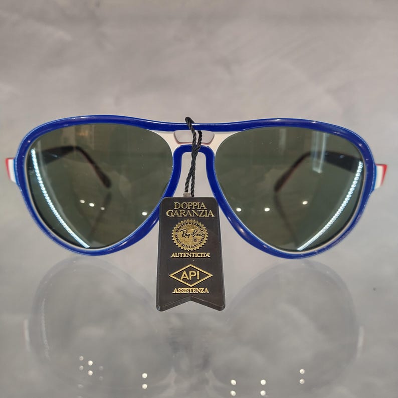 May include: Blue and white aviator sunglasses with green lenses. The sunglasses have a tag attached that says "Doppia Garanzia" and "Ray-Ban" with a logo. The tag also says "Autenticita" and "API Assistenza".
