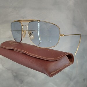 Puede incluir: Gafas de sol aviador doradas con lentes azules y una funda de cuero marr&oacute;n. Las gafas tienen una forma cuadrada y un fino marco de metal.