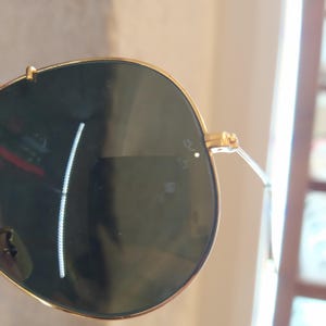 Puede incluir: Gafas de sol aviador doradas con lentes verde oscuro. Las gafas tienen un peque&ntilde;o punto blanco en la lente derecha.