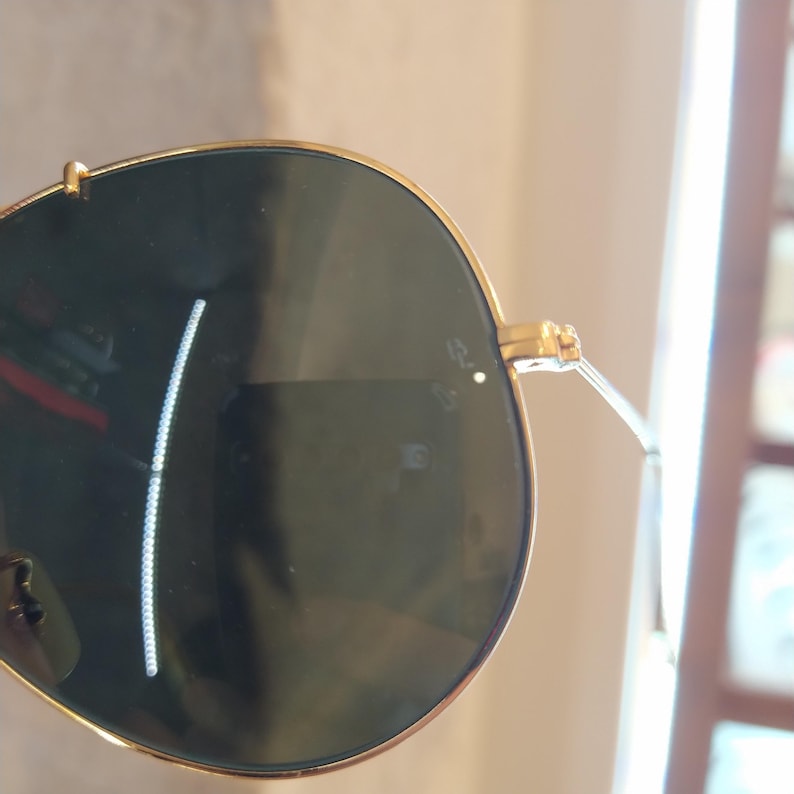 Puede incluir: Gafas de sol aviador doradas con lentes verde oscuro. Las gafas tienen un peque&ntilde;o punto blanco en la lente derecha.