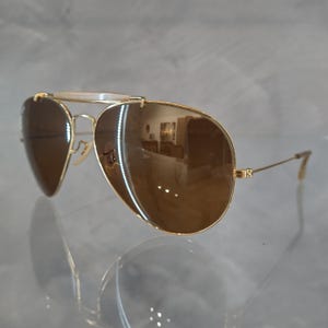 May include: Gold aviator sunglasses with brown lenses. The sunglasses have a thin metal frame and are reflected in the surface below.